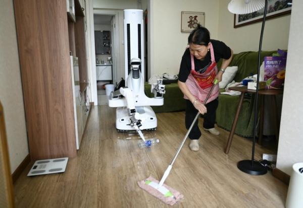 Un robot assistant ménager transporte une bouteille jusqu'à une poubelle tandis qu'une femme de ménage nettoie le sol chez une cliente à Pékin, le 21 mai 2026 en Chine