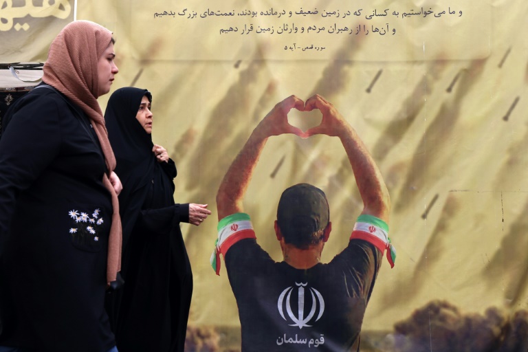 Des femmes passent devant une banderole représentant un homme faisant un cœur avec ses mains devant des tirs de missiles, à Téhéran le 19 avril 2026