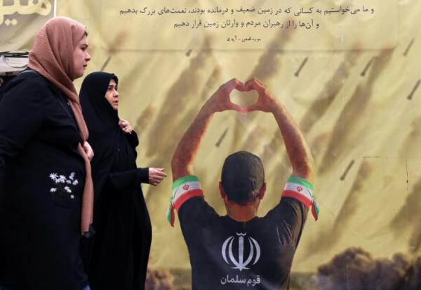 Des femmes passent devant une banderole représentant un homme faisant un cœur avec ses mains devant des tirs de missiles, à Téhéran le 19 avril 2026