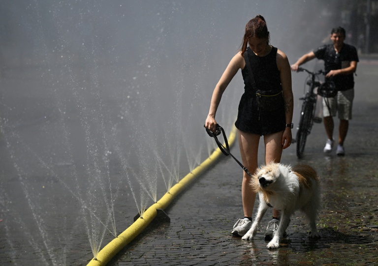 Une femme promène son chien devant des tuyaux d’arrosage déversant de l’eau pour apporter un peu de fraîcheur face à la chaleur estivale, dans le cadre des mesures d’adaptation climatique de Cologne, dans l’ouest de l’Allemagne, le 25 juin 2026